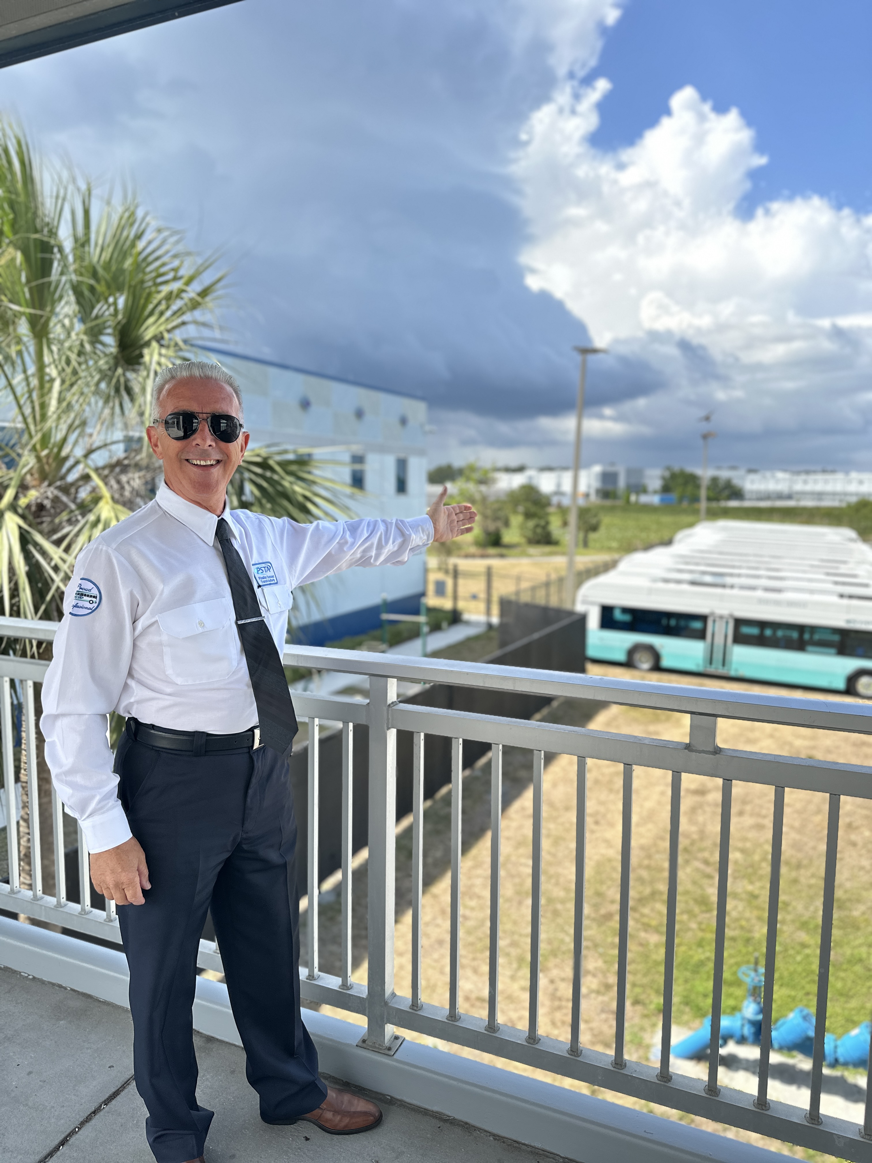 A photo of Milan wearing a white collared PSTA shirt, smiling and gesturing to a line of GILLIG electric buses.