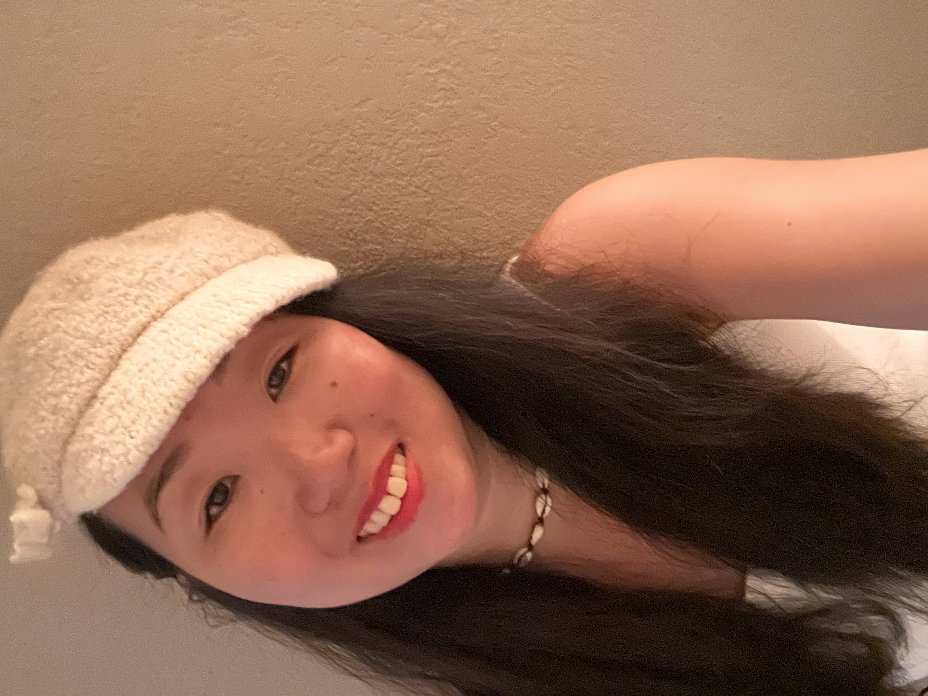 Selfie of Vanessa smiling, wearing a white tank top, white hat, and seashell necklace.