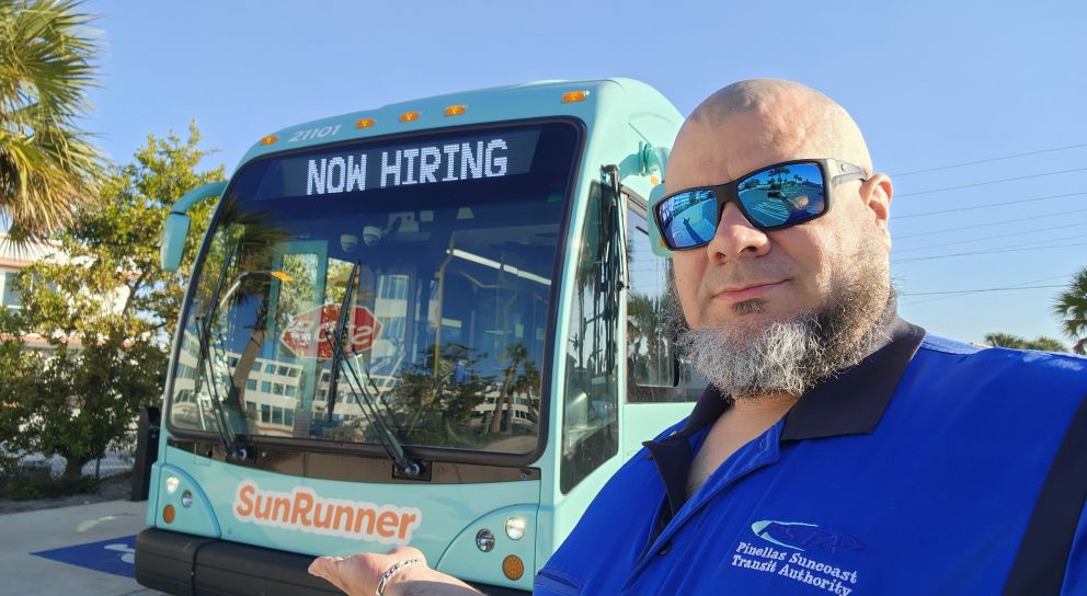 Brian standing in front of the SunRunner bus, holding his hand under the logo. The headsign on the bus says "Now Hiring." Brian wears a black and blue PSTA shirt and sunglasses.